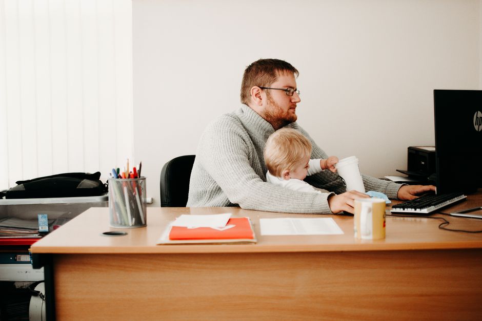 Un père jonglant avec son travail à son bureau tout en tenant son enfant, illustrant le télétravail et la parentalité.
