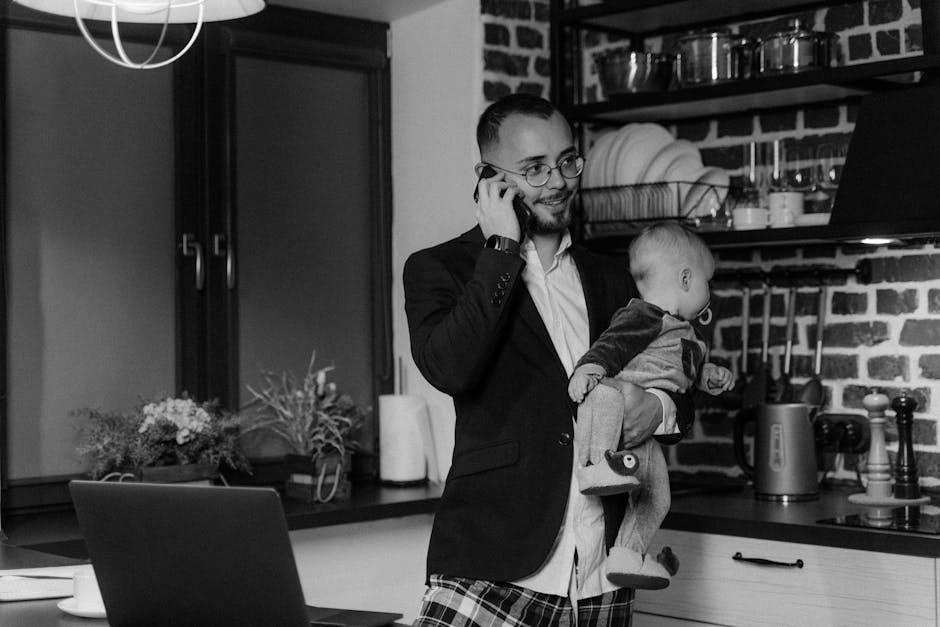 Un père jonglant avec son travail en tenant son bébé et en utilisant un téléphone dans une cuisine moderne.