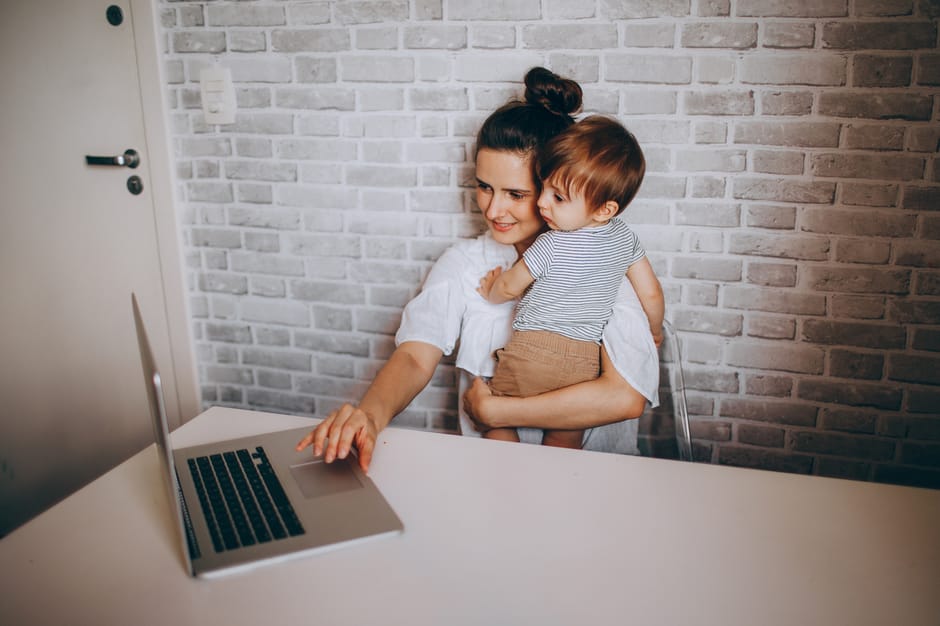 Vue de dessus d’une femme souriante regardant un ordinateur portable, assise à une table avec son jeune fils dans les bras et travaillant sur un projet à distance