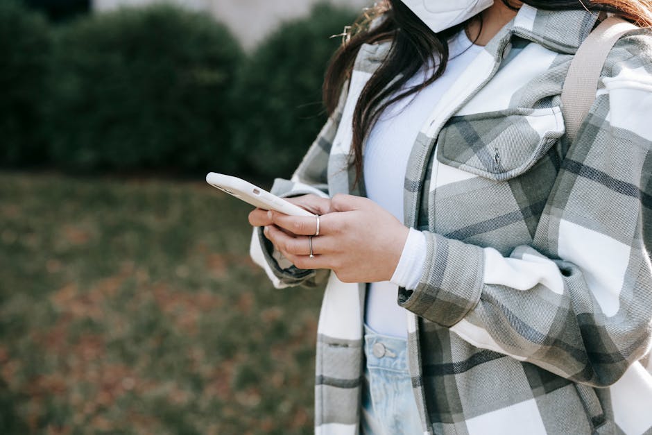 Crop anonymous young female in casual clothes and mask with backpack texting on smartphone in street in daylight
