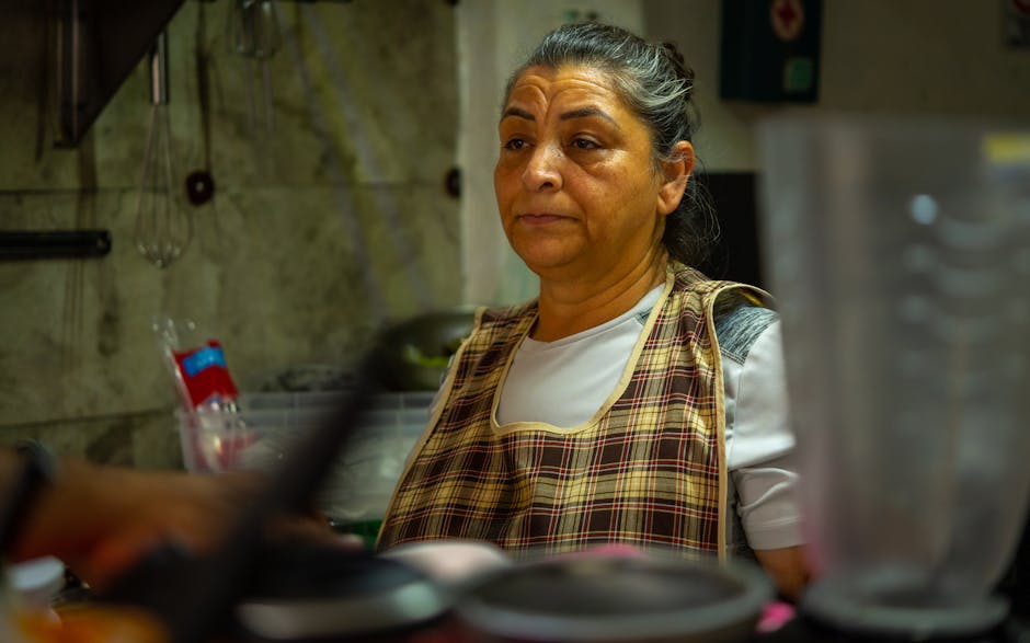 Une femme portant un tablier à carreaux se tient dans une cuisine, l’air pensif et peut-être fatigué