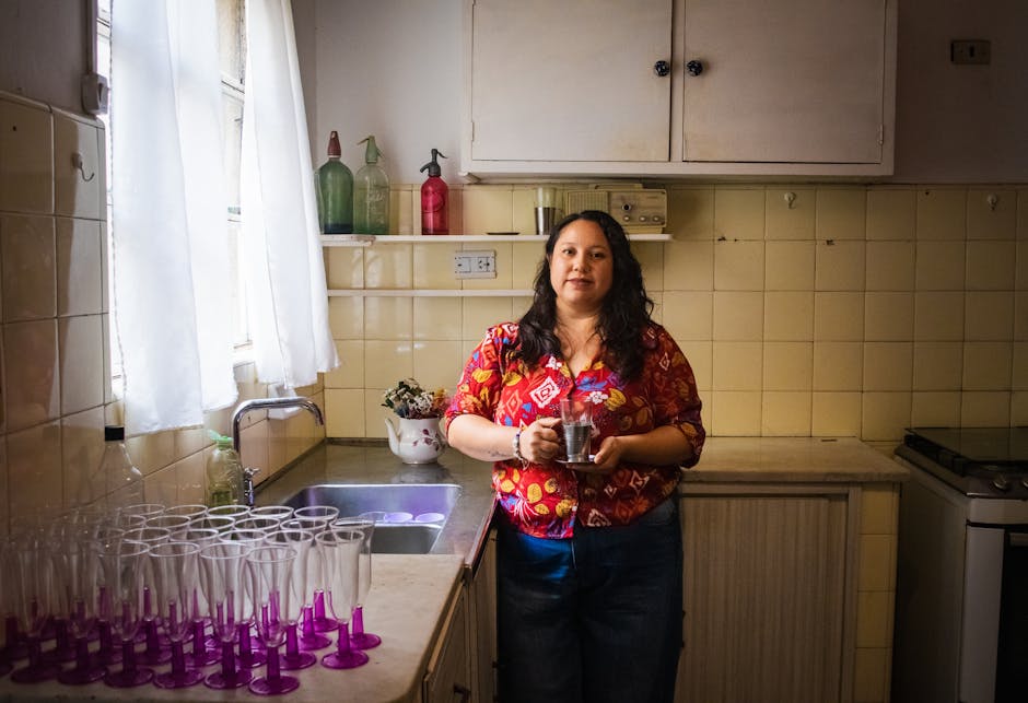 Une femme se tient dans une cuisine bien éclairée en tenant une tasse, entourée de verres et d’une décoration chaleureuse.