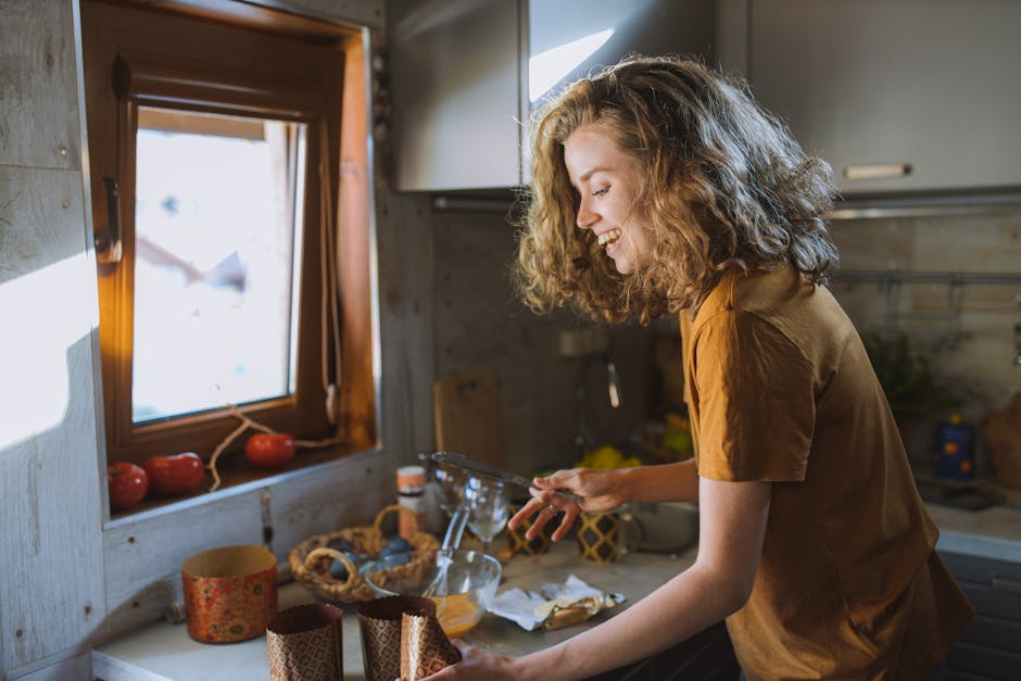 Une femme souriante aux cheveux bouclés prépare de la nourriture dans une cuisine ensoleillée.