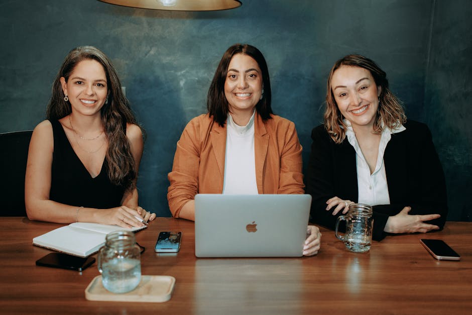 Tres mujeres profesionales sentadas en un escritorio de madera en una oficina con portátiles y cuadernos.