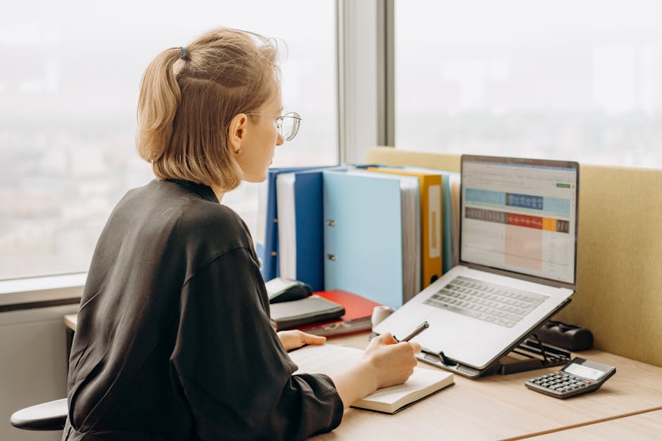 A side view of a professional woman working on a laptop with spreadsheets in a modern office.