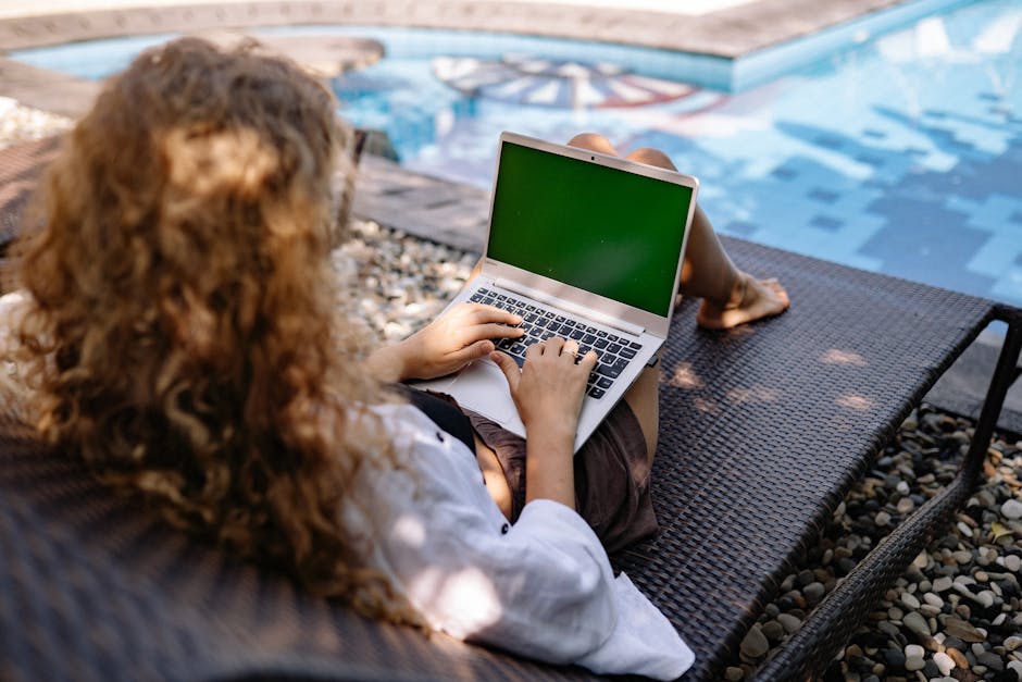 From above back view of anonymous female freelancer typing on portable computer while lying on sunbed near swimming pool with transparent water in sunlight