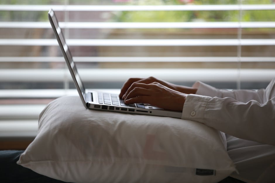 Close-up of hands typing on a laptop while seated on a pillow indoors.