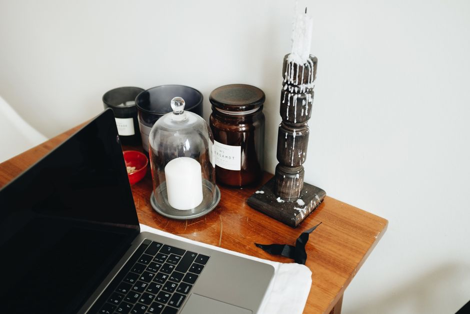 Freelance workspace with laptop and various black aromatic candles in glass jars and wooden candlestick placed on wooden table in light room of modern apartment