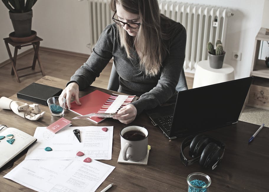 Woman working on design project at home office with laptop, papers, and color palette.