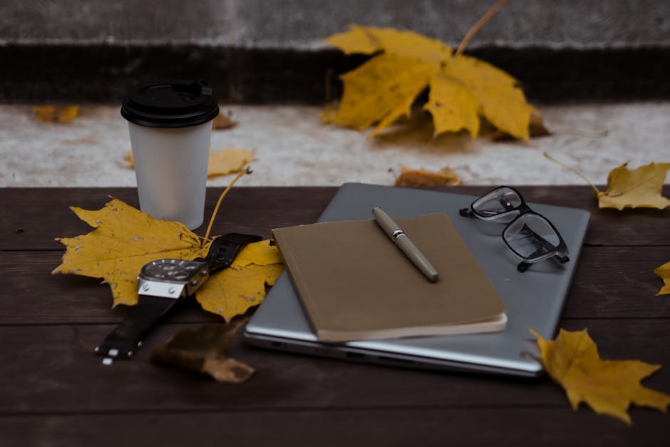 Cozy autumn workspace with coffee cup, notebook, and fallen leaves on wooden table.