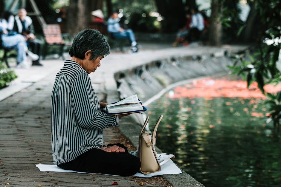Elderly woman enjoying a peaceful reading moment by a pond in a serene park setting.