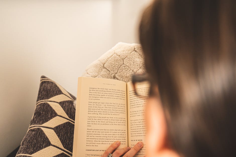 An adult reading a book in a cozy indoor setting with pillows and warm ambiance.