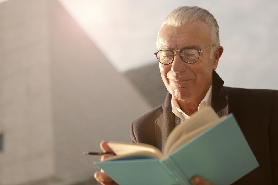 Elderly man wearing glasses, reading a book outdoors with a smile on a sunny day.