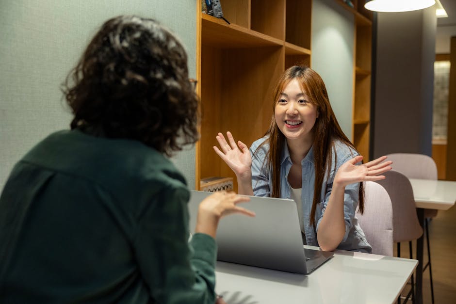 Two women having a cheerful work conversation at an office table with a laptop.