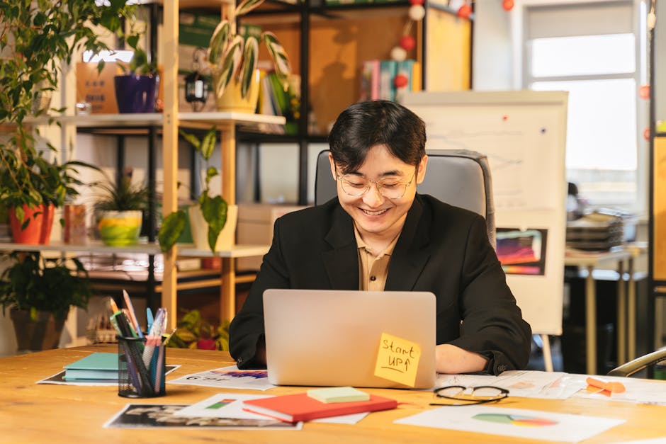 Smiling young professional in black suit working on laptop at a cozy office desk surrounded by plants.