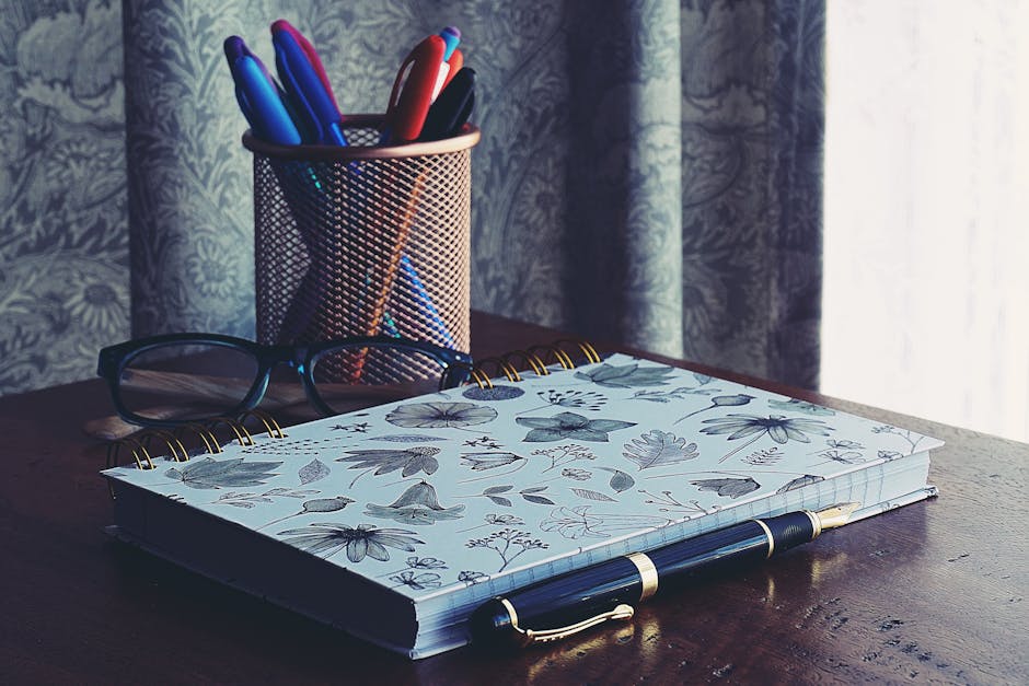 A stylish workspace featuring a floral notebook, glasses, and pens on a wooden desk.