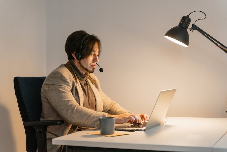 Professional man in a cozy office setting using a laptop for work-related tasks, wearing a headset under a desk lamp.